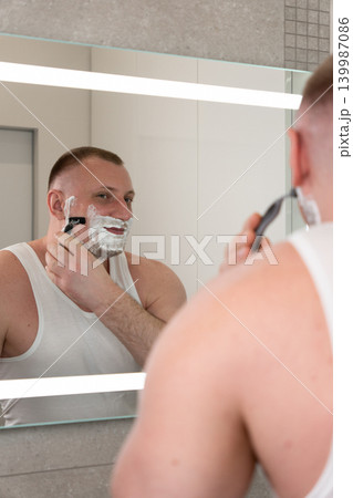 Vertical close up portrait of handsome young man shaving his face with razor in bathroom mirror during morning routine. Male grooming, skincare and personal care concept. 139987086