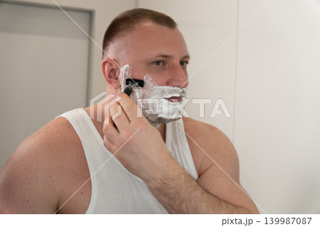 Handsome young man shaving his face with razor in bathroom mirror during morning routine. Male grooming, skincare and personal care concept. Handsome young man shaving his face with razor in bathroom mirror during morning routine. Male grooming, skincare and personal care concept. 139987087