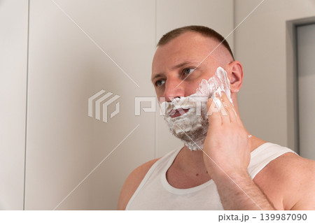 Close up portrait of handsome young man applying shaving foam on face in bathroom mirror. Morning male grooming and skincare routine concept before shaving. 139987090