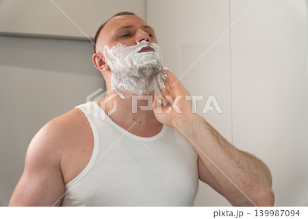 Close up portrait of handsome young man applying shaving foam on face in bathroom mirror. Morning male grooming and skincare routine concept before shaving. Close up portrait of handsome young man applying shaving foam on face in bathroom mirror. Morning male grooming and skincare routine concept before shaving. 139987094