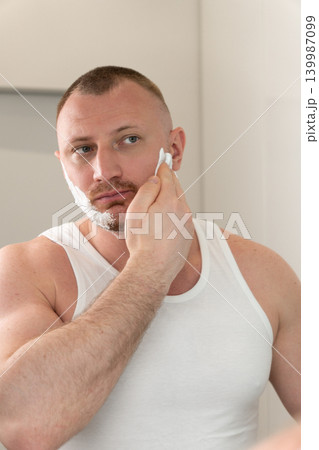 Vertical close up portrait of handsome young man applying shaving foam on face in bathroom mirror. Morning male grooming and skincare routine concept before shaving. 139987099