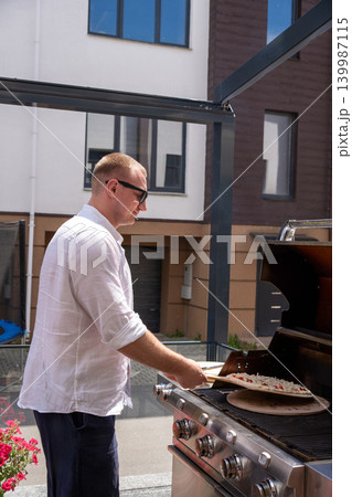 Vertical shot of a man preparing pizza on a gas grill on an outdoor terrace. The image captures a modern lifestyle cooking scene with focus on outdoor grilling, summer atmosphere, and homemade food 139987115