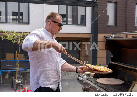 A man prepares pizza on a gas grill on a sunny outdoor terrace. The scene shows a modern lifestyle cooking moment with a focus on grilling food outdoors. Perfect for concepts of summer cooking A man prepares pizza on a gas grill on a sunny outdoor terrace. The scene shows a modern lifestyle cooking moment with a focus on grilling food outdoors. Perfect for concepts of summer cooking 139987120