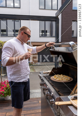 Vertical shot of a man preparing pizza on a gas grill on an outdoor terrace. The image captures a modern lifestyle cooking scene with focus on outdoor grilling, summer atmosphere, and homemade food Vertical shot of a man preparing pizza on a gas grill on an outdoor terrace. The image captures a modern lifestyle cooking scene with focus on outdoor grilling, summer atmosphere, and homemade food 139987123