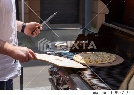 A man prepares pizza on a gas grill on a sunny outdoor terrace. The scene shows a modern lifestyle cooking moment with a focus on grilling food outdoors. Perfect for concepts of summer cooking A man prepares pizza on a gas grill on a sunny outdoor terrace. The scene shows a modern lifestyle cooking moment with a focus on grilling food outdoors. Perfect for concepts of summer cooking 139987125