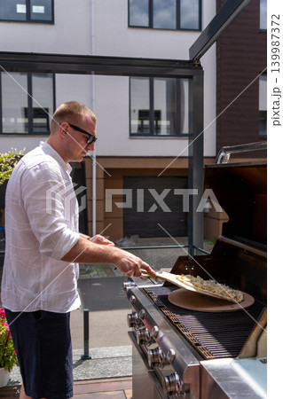 Vertical shot of a man preparing pizza on a gas grill on an outdoor terrace. The image captures a modern lifestyle cooking scene with focus on outdoor grilling, summer atmosphere, and homemade food Vertical shot of a man preparing pizza on a gas grill on an outdoor terrace. The image captures a modern lifestyle cooking scene with focus on outdoor grilling, summer atmosphere, and homemade food 139987372
