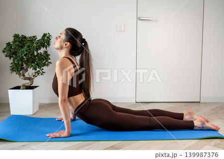 Young woman practicing stretching exercises with fitness resistance band at home. Female doing flexibility and mobility workout in cozy apartment interior. Healthy lifestyle, home fitness, wellness Young woman practicing stretching exercises with fitness resistance band at home. Female doing flexibility and mobility workout in cozy apartment interior. Healthy lifestyle, home fitness, wellness 139987376