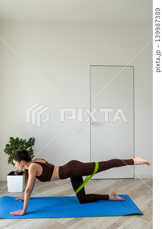 Vertical portrait of a fit young woman performing stretching and fitness exercises with resistance band at home. Female training indoors in sportswear during wellness and healthy lifestyle routine 139987389