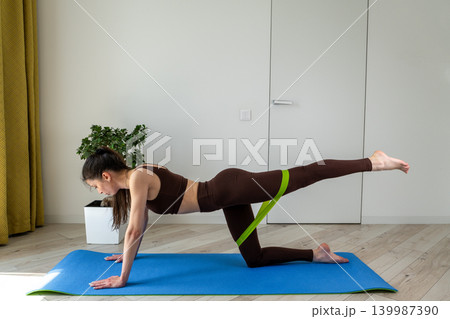 Young woman practicing stretching exercises with fitness resistance band at home. Female doing flexibility and mobility workout in cozy apartment interior. Healthy lifestyle, home fitness, wellness Young woman practicing stretching exercises with fitness resistance band at home. Female doing flexibility and mobility workout in cozy apartment interior. Healthy lifestyle, home fitness, wellness 139987390