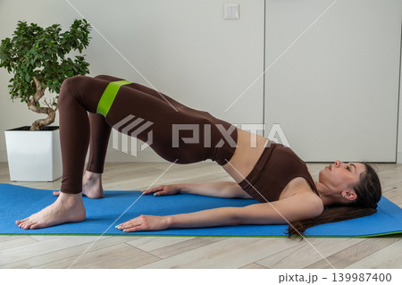 Young woman practicing stretching exercises with fitness resistance band at home. Female doing flexibility and mobility workout in cozy apartment interior. Healthy lifestyle, home fitness, wellness Young woman practicing stretching exercises with fitness resistance band at home. Female doing flexibility and mobility workout in cozy apartment interior. Healthy lifestyle, home fitness, wellness 139987400