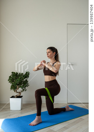 Vertical portrait of a fit young woman performing stretching and fitness exercises with resistance band at home. Female training indoors in sportswear during wellness and healthy lifestyle routine Vertical portrait of a fit young woman performing stretching and fitness exercises with resistance band at home. Female training indoors in sportswear during wellness and healthy lifestyle routine 139987404