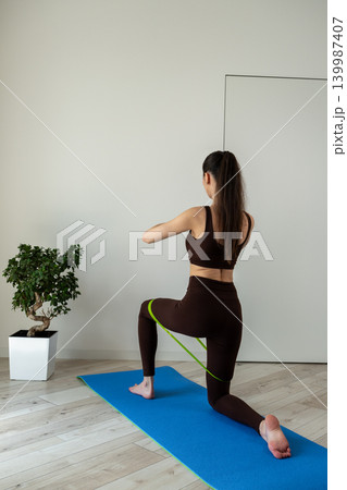 Vertical portrait of a fit young woman performing stretching and fitness exercises with resistance band at home. Female training indoors in sportswear during wellness and healthy lifestyle routine Vertical portrait of a fit young woman performing stretching and fitness exercises with resistance band at home. Female training indoors in sportswear during wellness and healthy lifestyle routine 139987407