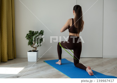 Young woman practicing stretching exercises with fitness resistance band at home. Female doing flexibility and mobility workout in cozy apartment interior. Healthy lifestyle, home fitness, wellness Young woman practicing stretching exercises with fitness resistance band at home. Female doing flexibility and mobility workout in cozy apartment interior. Healthy lifestyle, home fitness, wellness 139987417
