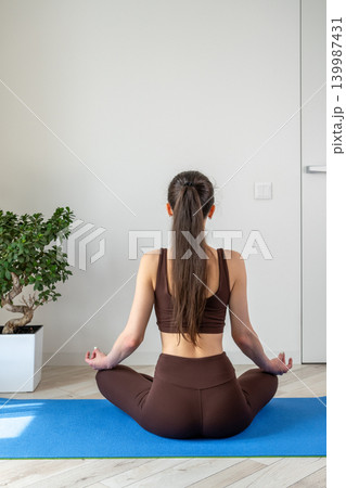 Vertical portrait of a peaceful young woman meditating and practicing yoga indoors. Female sitting cross legged on a yoga mat in a cozy apartment during morning wellness routine. Concept of 139987431
