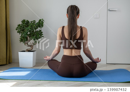 Young woman meditating and practicing yoga at home in a cozy living room. Calm female sitting on yoga mat in lotus pose during morning wellness routine. Concept of mindfulness, healthy lifestyle 139987432