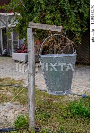 Old metal bucket hanging on a rustic wooden post in a countryside setting during cloudy weather Old metal bucket hanging on a rustic wooden post in a countryside setting during cloudy weather 139988170