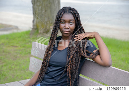Relaxed young African American woman with long braids sitting on a park bench looking at the camera Relaxed young African American woman with long braids sitting on a park bench looking at the camera 139989426