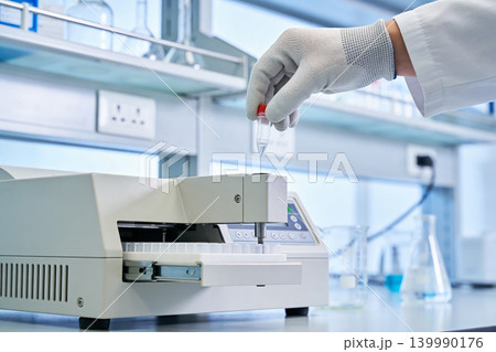 Scientist in lab coat and gloves carefully handles a small vial over automated equipment Scientist in lab coat and gloves carefully handles a small vial over automated equipment 139990176