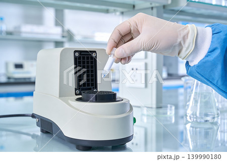 Scientist in lab coat and gloves places sample tube into laboratory heating device 139990180