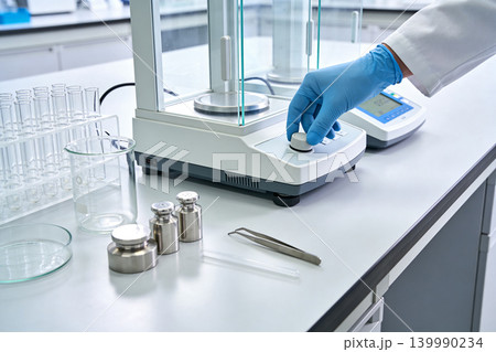 Scientist's gloved hand calibrating precise laboratory scale with weights and glassware 139990234