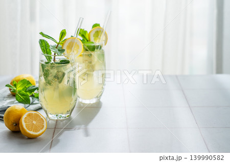 A summer drink with lemon, mint, and ice in a two glasses against a sunny window background. A summer drink with lemon, mint, and ice in a two glasses against a sunny window background. 139990582
