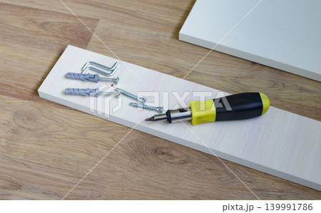 Screwdriver with a black and yellow plastic handle, plastic dowels, screws, and furniture boards on the floor, ready for furniture assembly. 139991786
