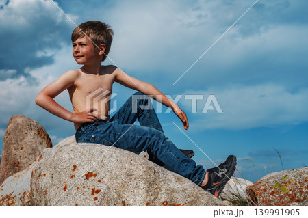 A seven-year-old boy in jeans sits on a boulder in the mountains A seven-year-old boy in jeans sits on a boulder in the mountains 139991905