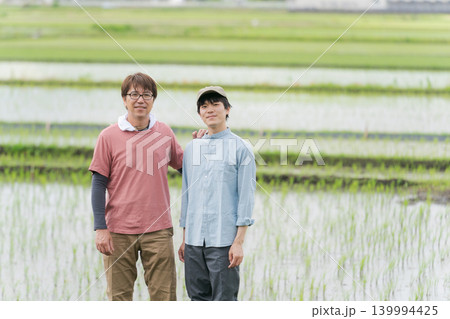田んぼの畦道に立つ20代と50代の農家 139994425