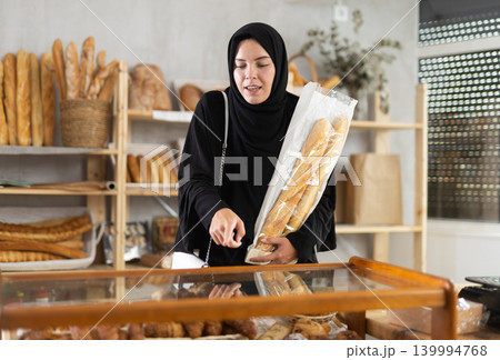 Girl in traditional Muslim clothing with a bag of baguettes in her hands chooses hot croissants in oriental bakery Girl in traditional Muslim clothing with a bag of baguettes in her hands chooses hot croissants in oriental bakery 139994768