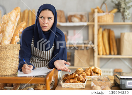 European woman in hijab reviewing paperwork while managing bakery 139994820