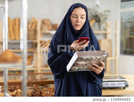 European Muslim woman in hijab customer at bakery scans QR code on cookie package using phone camera European Muslim woman in hijab customer at bakery scans QR code on cookie package using phone camera 139994993