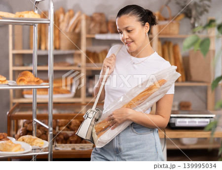 Asian woman buyer with baguettes in plastic package stands in trading floor of bakery Asian woman buyer with baguettes in plastic package stands in trading floor of bakery 139995034