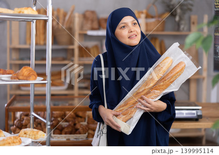 Asian Muslim woman buyer with baguettes in plastic package stands in trading floor of bakery 139995114