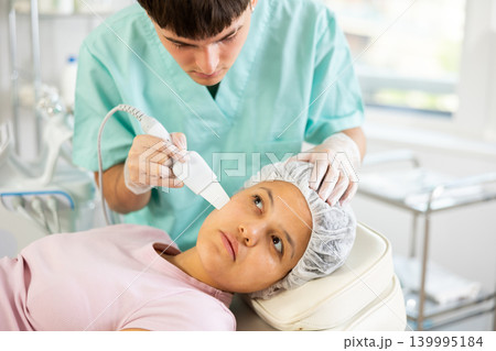Young male cosmetologist doing exfoliating procedure to young woman Young male cosmetologist doing exfoliating procedure to young woman 139995184