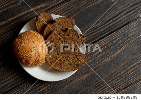 Sesame seed bun and several slices of dark, rustic rye bread on white plate on textured, dark brown wooden table, copy space. 139998209