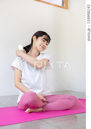 ヨガマットの上でマッサージガンで肩をセルフマッサージする日本人の若い女性 139998242
