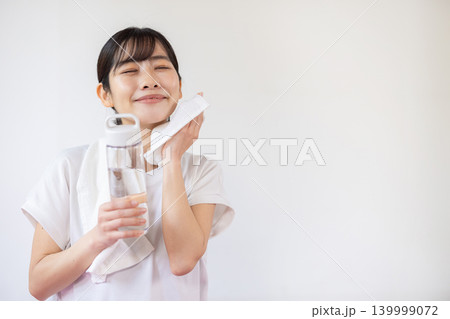 明るい室内でウォーターボトルを手に持ち、タオルで汗を拭きながら微笑む日本人の若い女性 明るい室内でウォーターボトルを手に持ち、タオルで汗を拭きながら微笑む日本人の若い女性 139999072