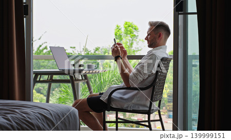 Man Using Smartphone At Hotel Balcony Workspace During Remote Work Routine, Digital Nomad Lifestyle And Mobile Productivity Concept Man Using Smartphone At Hotel Balcony Workspace During Remote Work Routine, Digital Nomad Lifestyle And Mobile Productivity Concept 139999151