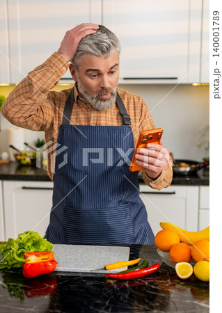 Middle-aged man shocked by bad news on smartphone in home kitchen after sudden internet message loss 140001789