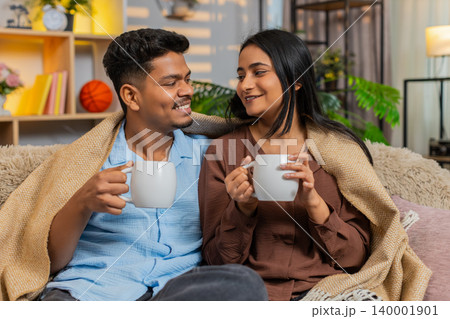 Indian couple man and woman sitting on home sofa with tea under blanket enjoying winter home comfort Indian couple man and woman sitting on home sofa with tea under blanket enjoying winter home comfort 140001901