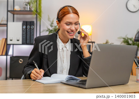 Young businesswoman wears headset and takes notes during client video call on laptop smiling calmly Young businesswoman wears headset and takes notes during client video call on laptop smiling calmly 140001903