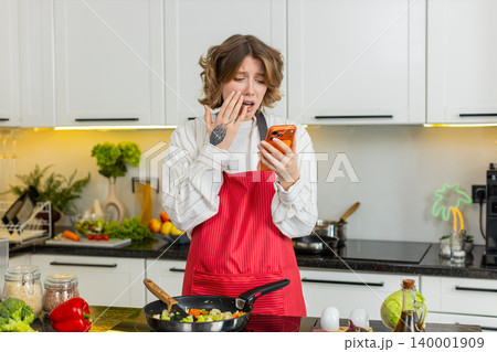 Young woman upset after bad smartphone message during home cooking feeling loss failure and defeat Young woman upset after bad smartphone message during home cooking feeling loss failure and defeat 140001909