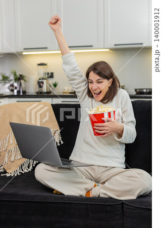 Young woman cheers match on laptop jumps at goal claps shows thumbs up and eats popcorn during game Young woman cheers match on laptop jumps at goal claps shows thumbs up and eats popcorn during game 140001912
