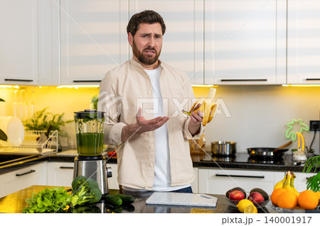 Middle-aged man confused holds banana near blender choosing healthy snack in kitchen at home concept Middle-aged man confused holds banana near blender choosing healthy snack in kitchen at home concept 140001917