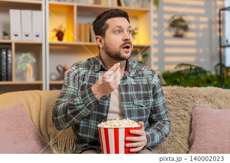 Young man on home sofa watching movie with popcorn and reacting with surprise to exciting film scene Young man on home sofa watching movie with popcorn and reacting with surprise to exciting film scene 140002023
