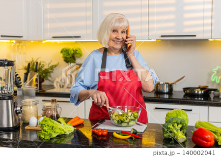 Senior old woman mixing vegetable salad while talking on phone in home kitchen for healthy diet plan 140002041