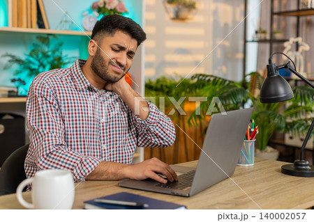 Indian man on home table working on laptop and touching neck with pain during remote work shift mode Indian man on home table working on laptop and touching neck with pain during remote work shift mode 140002057