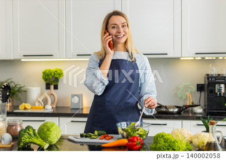 Young woman talking on orange smartphone while mixing salad in kitchen sharing healthy keto recipe Young woman talking on orange smartphone while mixing salad in kitchen sharing healthy keto recipe 140002058