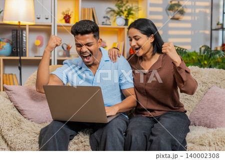 Indian couple man and woman sitting on home sofa celebrating approval on laptop with joyful victory 140002308