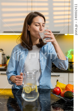Young woman pouring cold lemon water in home kitchen to quench thirst for diet hydration fit routine Young woman pouring cold lemon water in home kitchen to quench thirst for diet hydration fit routine 140002411